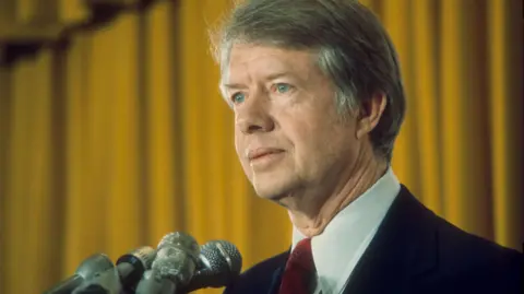 Jimmy Carter stands in front of a gold curtain, wearing a black blazer and a red tie, stood by a cluster of microphones.