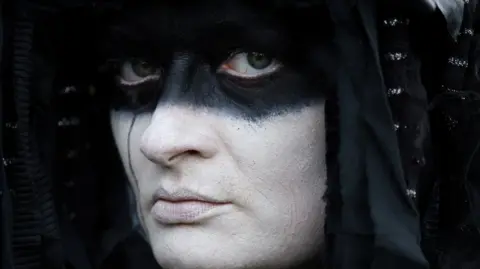 Reuters An activist of "Extinction Rebellion" (XR) dressed as an oil slick, attends a protest, in Windsor, Britain, August 30, 2024. The activist has a painted white face with a black around the eyes and wears a black hood.