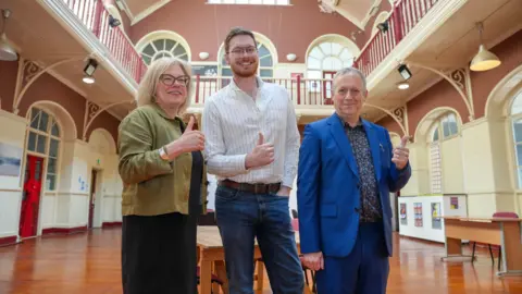 A man and two women stand in a line in a hall and smile at the camera, each of them doing a thumbs up.