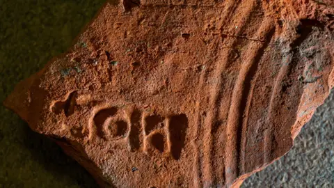 Tony Jolliffe/BBC A red roof tile has a stamp made up of three letters on its surface showing that it was a public building and belonged to the procurator of London. It also has a series of curved grooves made by the tile maker dragging his fingers across it when it was being produced.