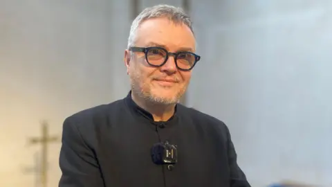 Jenny Kirk/BBC Chris Sanham, Church Verger dressed in all black and has short grey hair and is wearing dark rimmed circular glasses.