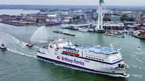 Brittany Ferries The Barfleur leaving Portsmouth Harbour with tugs following behind and the shoreline of Portsmouth in the background on an overcast day