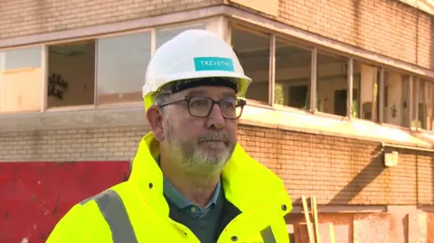 Man standing in front of derelict building.  He is wearing high visibility clothing and a hard hat. 