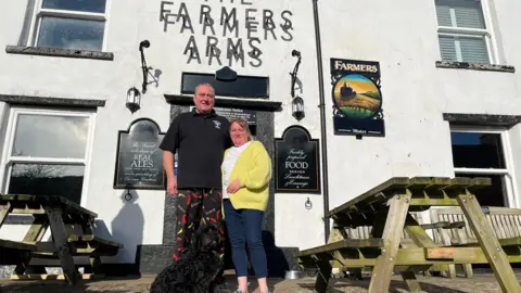 A man wearing a black top and patterned trousers is standing next to a woman with a yellow cardigan, white shirt and blue jeans.  
Sitting in front of  them is a black spoodle dog.
They are all in front of a white building with a sign saying "Farmers Arms" and there are wooden picnic benches either side. 