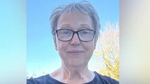Christine Grant is seen from her shoulders up. She is wearing a dark blue T-Shirt and has short, grey hair. The sky behind her is blue.