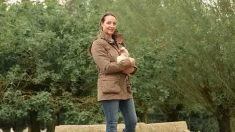 St. Ewe Free Range Eggs Rebecca Tonks stood on a wooden trailer holding a chicken. There are trees in the background. She's wearing a brown coat, and the chicken is white with a red face. 