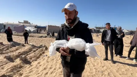 BBC A man in a cap carries a small bag wrapped in cloth containing the body of his newborn baby