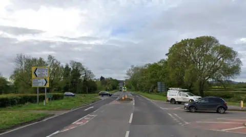 Google Lock's Garage junction shows the main A465 road, with roads going off in both directions to Abergavenny and Hay-on-Wye. There are white lines and chevron road markings and give way lines, as well as a bollard in the middle. Cars are crossing the junction in the Google Maps image.