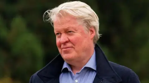 A man with light-coloured hair wearing a dark coat over a light blue button-up shirt, photographed outdoors with blurred green foliage in the background.
