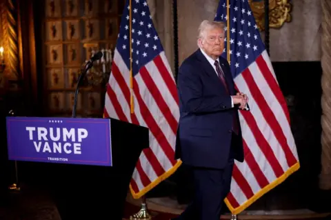 Reuters US President-elect Donald Trump stand by a lectern with the words "Trump-Vance transition". Two US flags are position behind Trump, who wears a dark suit and purple tie.