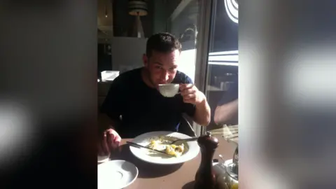 Facebook A young man with short dark hair and stubble drinks a cup of tea and eats a cooked breakfast in a cafe. 