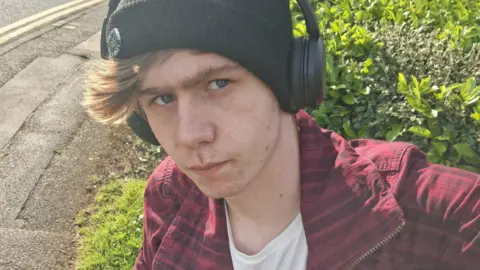 Clover A close-up image of a young man's face. He is frowning, wearing a close-fitting black woollen hat, headphones and a red zip-jacket over a white t-shirt.