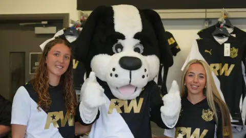 Port Vale Football Club A dog mascot poses between two young women in matching black and white football jerseys. The women are smiling and there are shirts in the background.

