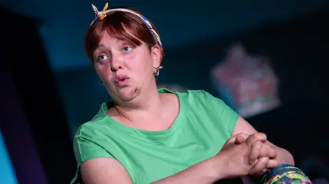 Bernie McCallister Caroline Curran, the actress in the role of Ciara. She is a woman with fair to brown hair, with a hair band. She is wearing a green t-shirt. 