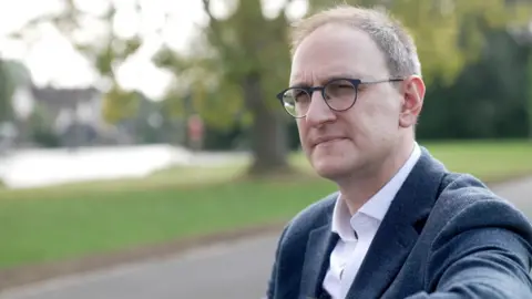 Ant Saddington/BBC Ian Sollom MP is sat, looking to the side. He's wearing glasses and a suit