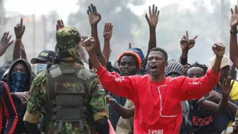A group of protesters - one in a red top clearly seen at the front - hold their arms in the air as they face a policeman in camouflage uniform.