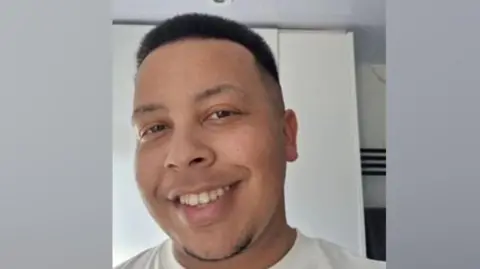 Family photograph Dominic Lloyd smiles wearing a white t-shirt while stood indoor against a white wall. 