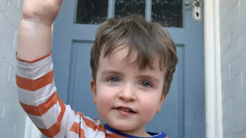 Dan Booth Charlie is sat down by a front door waving. He has an orange stripy top on. He has short brown hair.