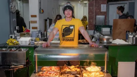 A man wearing a yellow t-shirt which says 'Lifeshare' stands behind a bain-marie with hot foot in it. 