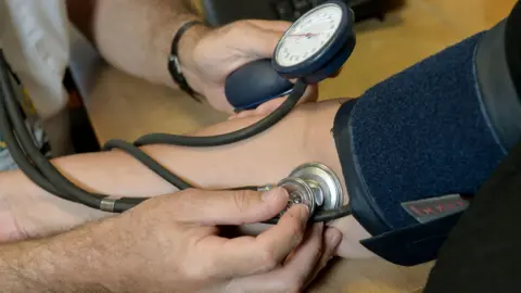 A person is having their blood pressure taken. They are wearing a cuff on their right arm and a doctor is holding the stethoscope against their arm. The shot is a close-up and faces are not visible.