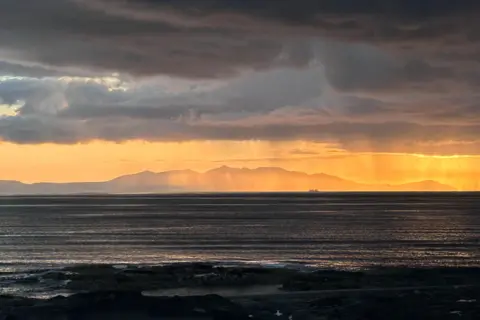 David Smith A ferry in the far distance, silhouetted against hills, under an orange sunset sky.