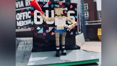 Brad Charlton-Wells A small lego build of a girl with brown hair wearing black boots, blue shorts and a white crop top. She is holding a microphone and a red megaphone. Behind her is a black screen with colourful shape on it and the word GUTS in white bricks.