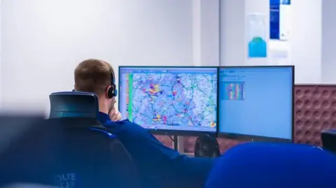 Cambridgeshire Police A police call handler can be seen from behind in an office. He is wearing a blue uniform and has short brown hair and a beard. He has headphones on his head and there are two computer screens in front of him, one showing a map.