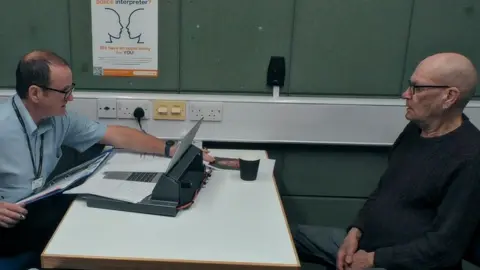 The Garden Productions Two men in a police interview room, across a white table from one another. On the left is an officer wearing a light blue shirt and glasses leaning over a white table and pointing to a photograph. On the right is David Newton, wearing dark framed glasses and a dark blue jumper. 