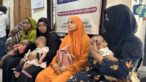 Several women with babies sit closely together on a bench inside a hospital waiting area. They are wearing black, green and orange outfits. A poster with Bengali text about vaccination is visible on the wall behind them, along with a doorway and other clinic signage.