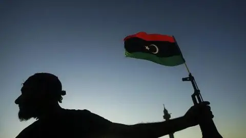 An anonymous Libyan fighter, in shadow, holds a rifle with a Libyan flag attached it flying in the air
