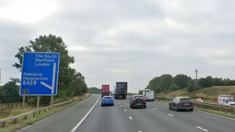Cars and trucks on a three-lane motorway. On the left is a blue sign showing directions for The South, Sheffield, London, Barnsley and Manchester