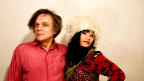 Artist Sergei Ivanov wearing a bright textured shirt stands beside his partner Tszman Chan dressed in patterned clothing. She is wearing a large, fluffy light‑coloured hat and has a scarf draped across the shoulder. Both individuals pose against a plain, softly lit background.