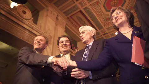 Getty/Hulton Archive/Peter MacDiarmid The SDLP team celebrating after ministerial positions were agreed on 29 November 1999.  Sean Farren is shaking hands with Bríd Rodgers. They are standing at either side of John Hume and Seamus Mallon in the Great Hall in Stormont. 