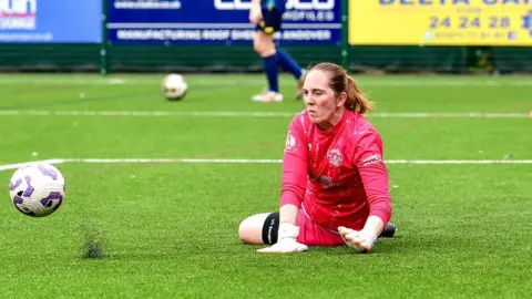 Holly Robinson making a save on a 3G pitch, with rubber pellets bouncing off the pitch.