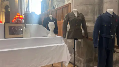 A small table is draped in a white cloth with the head of a mannequin, a framed black and white photo and four mannequins displaying army uniforms and overalls are on display at the church.