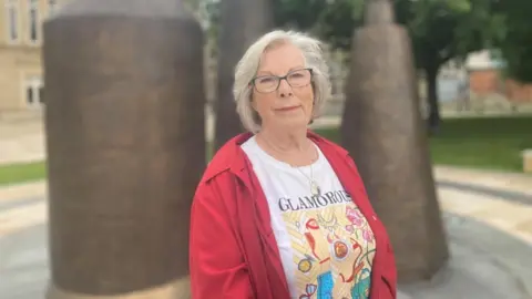 Clare is looking at the camera. The photo has been taken from the waist up. She has a printed t-shirt on that is mostly white with a yellow pattern, the word glamorous is at the top. She is wearing a red rain jacket and has short grey hair. She is also wearing black framed glasses. 