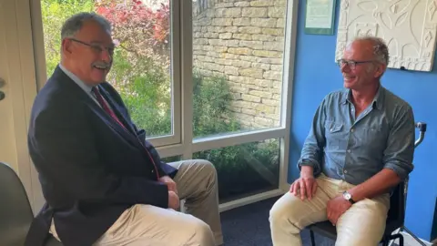 Jonathan Pape and Dr Simon Opher MBE. Both men are sat on chairs facing each other. They are pictured smiling.