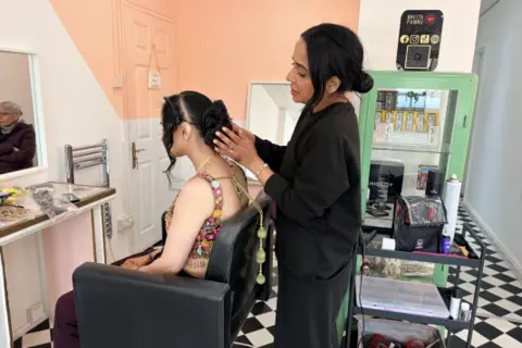 A woman in a black sweater and trousers stands in a salon with black and white checked floors. She stands behind a woman sat in a black salon chair who has her hair in a bun. The woman standing touches the other woman's hair.