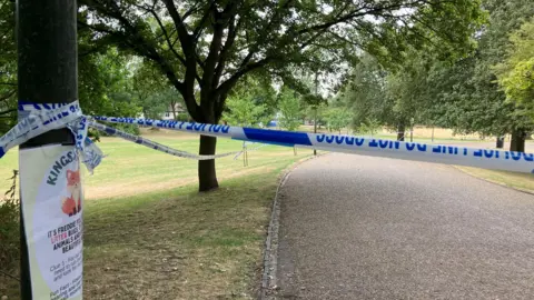 Blue and white police tape across a wide path in parkland. The tape runs between trees with grassy areas underneath, next to a wide path.