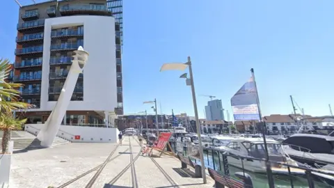 Blocks of flats around a marina with lines of boats on the water to the right and a wide path with benches overlooking the water alongside.