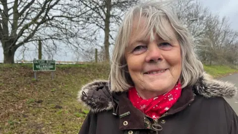 Penny Carpenter smiling at the camera, she is wearing a brown jacket and red scarf stood by a rural road sign and tree