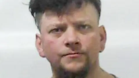 A police picture of a man with dark hair with a floppy fringe and a light goatee beard staring straight into the camera
