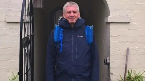 Handout photo of Ian Evans. He is stood in a doorway wearing a navy blue coat. He is looking directly at the camera and smiling.