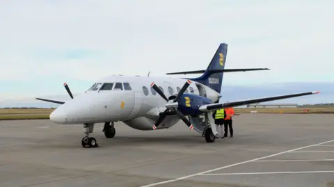 A Jetstream J32 aircraft sat on the runway. It's a grey plane with navy blue and yellow wings and black propellers with red and white tips.