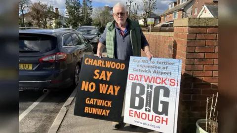 A man with short white hair and glasses wearing a navy jumper under a green gilet. He is looking at the camera while standing on the street holding two signs. One says "Charlwood say no way Gatwick runway", and the other says "Say no to further expansion of Gatwick Airport. Gatwick's big enough".