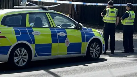 A police car and two officers stood next to police tape in a residential area. 