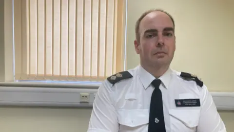 Jon Belcher is sat at his desk in uniform with a window with blinds behind him. He is looking at the camera, with a fairly serious expression. He has short brown hair.