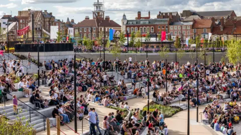 Stockton Council Dozens of people line the steps of Stockton Waterfront's amphitheatre. Brown brick buildings on the high street are visible in the background.