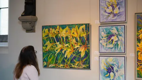 Ushaw A woman with long brown hair looking at a painting on the wall. It is a large painting with a repeated flower pattern with yellow, orange and green colours. Next to that painting are three similar ones stacked on top of each other, but purple is the main colour. They are set on a white wall.