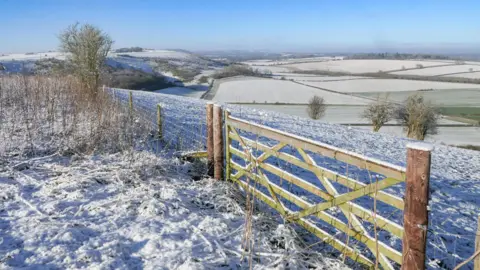 BobandMarleyDog A snowy countryside scene. There are rolling fields covered in snow stretching to the horizon under a blue sky. In the foreground a wooden gate is topped with snow.